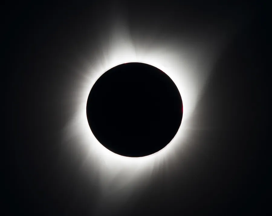The Sun's white corona streaming outwards around the black disc of the Moon, with two small pink prominences at the edge, against a dark sky.
