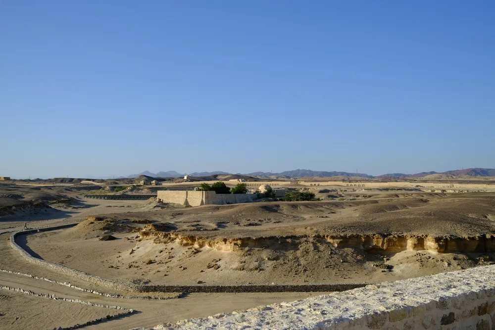 Arid coastal desert at Marsa Alam: low stone buildings on a plain of pale sand and rock, bare hills on the horizon under a cloudless sky.