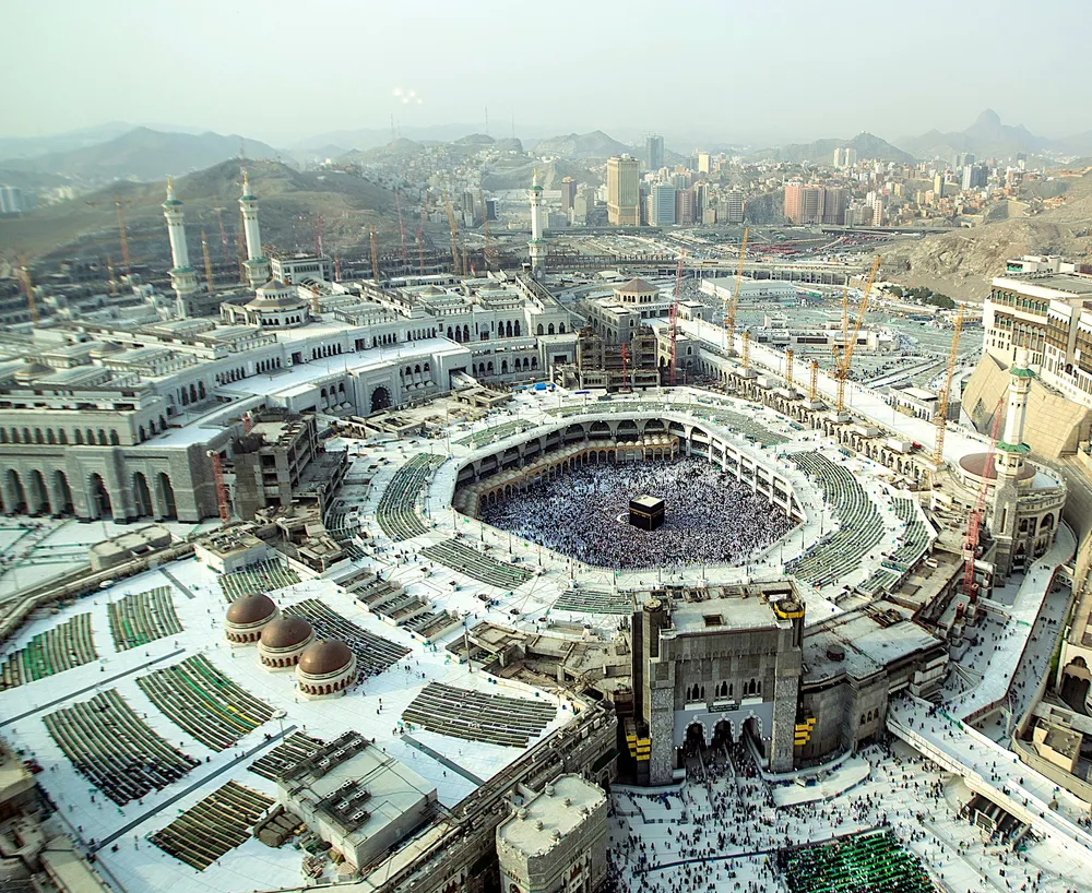 Aerial view of the Grand Mosque of Mecca: the black cube of the Kaaba at the centre of a circular courtyard packed with worshippers, ringed by the mosque and its galleries, with construction cranes around it and the buildings and hills of Mecca behind.